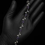 A silver vine bracelet with purple gemstones and clear crystal leaves, displayed on a hand wearing a black glove against a black background.