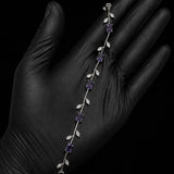 A silver vine bracelet with purple gemstones and clear crystal leaves, displayed on a hand wearing a black glove against a black background.