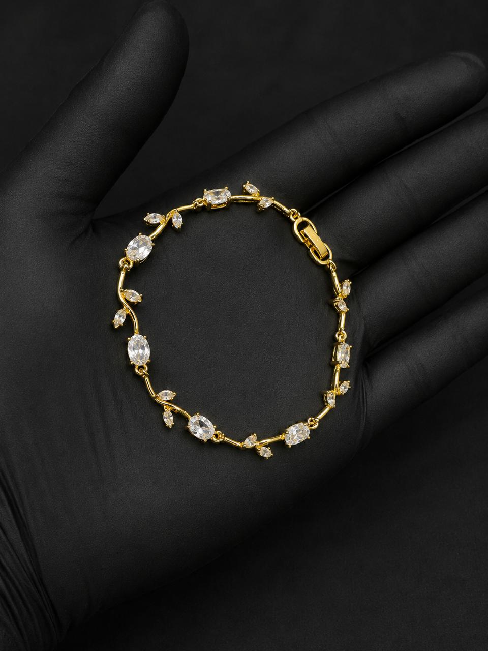 A delicate gold bracelet with clear gemstones arranged in a vine pattern, displayed on a black gloved hand against a dark background.