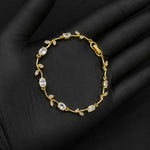A delicate gold bracelet with clear gemstones arranged in a vine pattern, displayed on a black gloved hand against a dark background.
