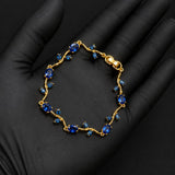 A gold bracelet with blue gemstones, displayed on a black gloved hand against a dark background.