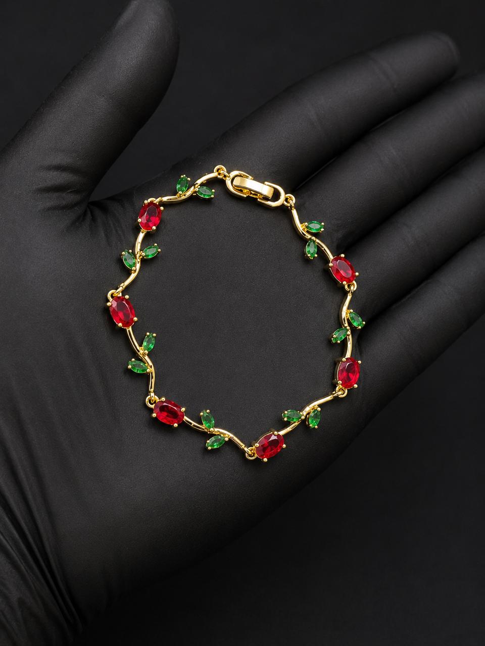 A gold bracelet with red oval gemstones and small green leaf accents, displayed on a black gloved hand.