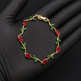 A gold bracelet with red oval gemstones and small green leaf accents, displayed on a black gloved hand.