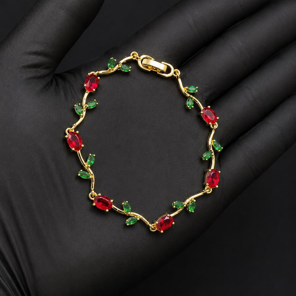 A gold bracelet with red oval gemstones and small green leaf accents, displayed on a black gloved hand.