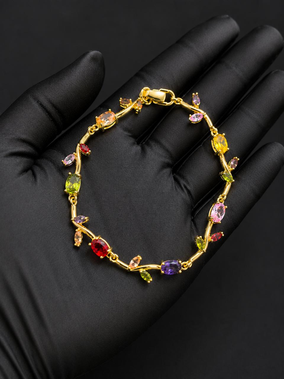 A gold bracelet with colorful gemstones displayed on a black gloved hand. The bracelet features a gold vine-like design with multiple colored stones in red, yellow, green, purple, and pink.