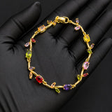 A gold bracelet with colorful gemstones displayed on a black gloved hand. The bracelet features a gold vine-like design with multiple colored stones in red, yellow, green, purple, and pink.