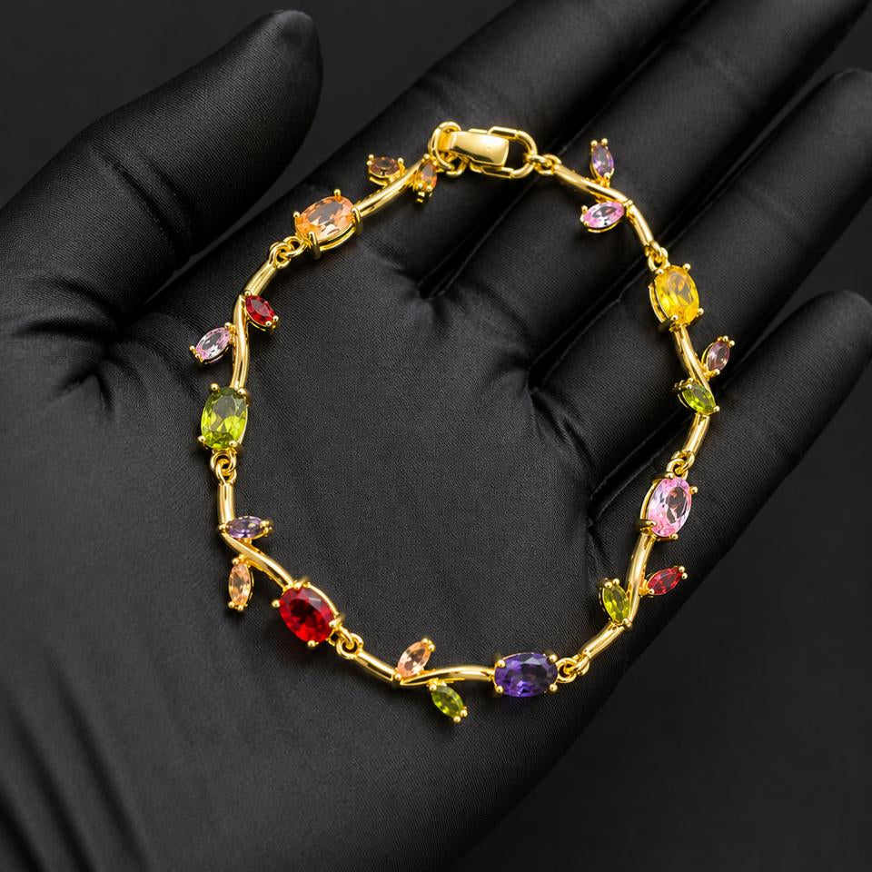 A gold bracelet with colorful gemstones displayed on a black gloved hand. The bracelet features a gold vine-like design with multiple colored stones in red, yellow, green, purple, and pink.