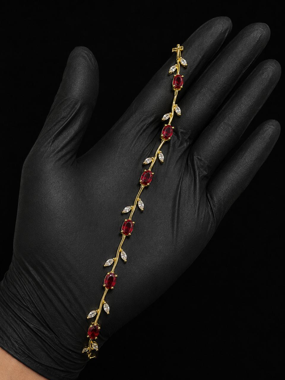 A gold vine-style bracelet with red gemstones and clear stones, displayed on a hand wearing a black glove against a black background.