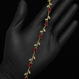 A gold vine-style bracelet with red gemstones and clear stones, displayed on a hand wearing a black glove against a black background.