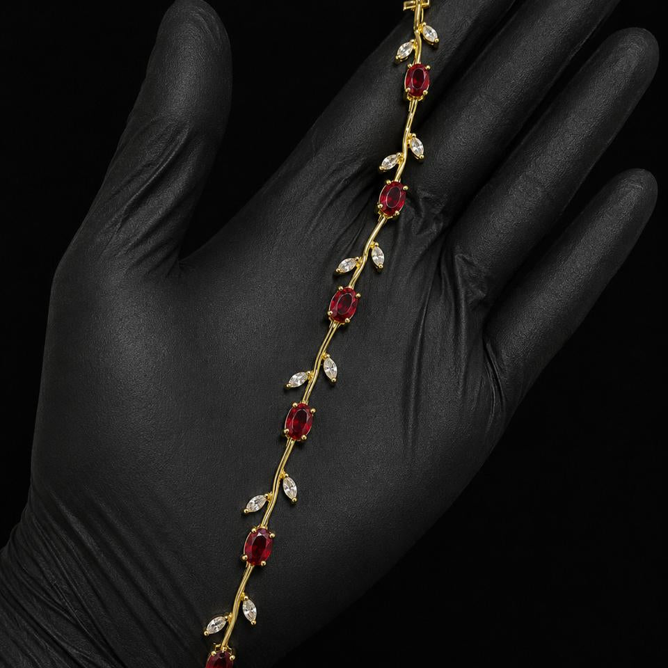 A gold vine-style bracelet with red gemstones and clear stones, displayed on a hand wearing a black glove against a black background.