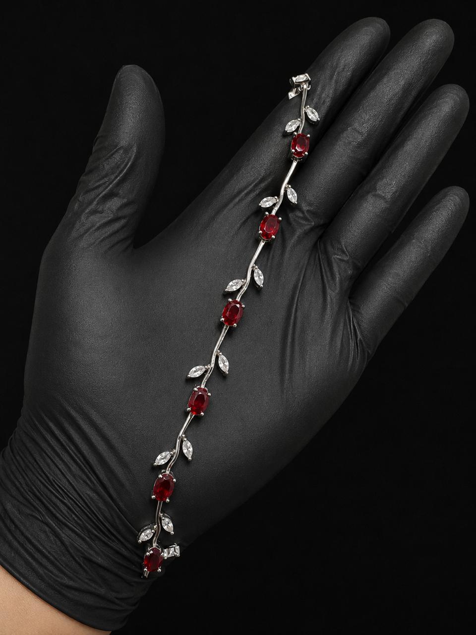 A silver vine bracelet with red gemstone flowers and clear crystal leaves, displayed on a hand wearing a black nitrile glove against a black background.