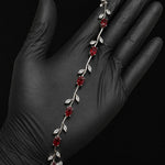 A silver vine bracelet with red gemstone flowers and clear crystal leaves, displayed on a hand wearing a black nitrile glove against a black background.