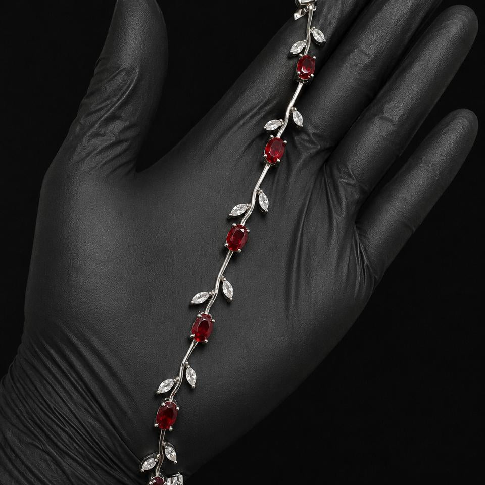 A silver vine bracelet with red gemstone flowers and clear crystal leaves, displayed on a hand wearing a black nitrile glove against a black background.