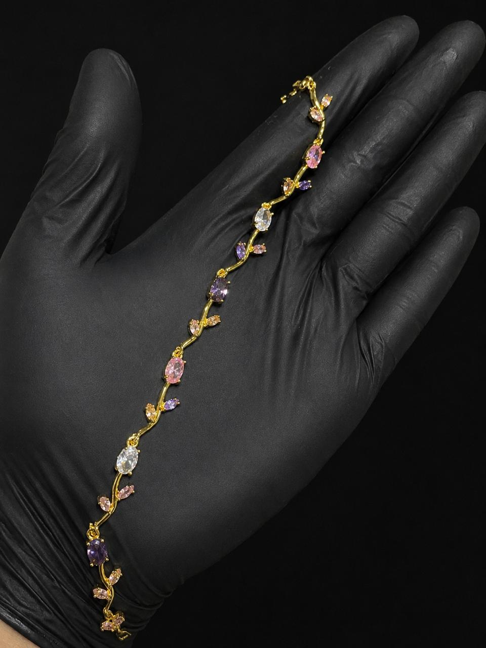 A delicate gold vine-style bracelet with pink, purple, and clear gemstones, displayed on a hand wearing a black glove against a solid black background.