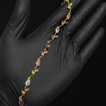 A delicate gold vine-style bracelet with pink, purple, and clear gemstones, displayed on a hand wearing a black glove against a solid black background.