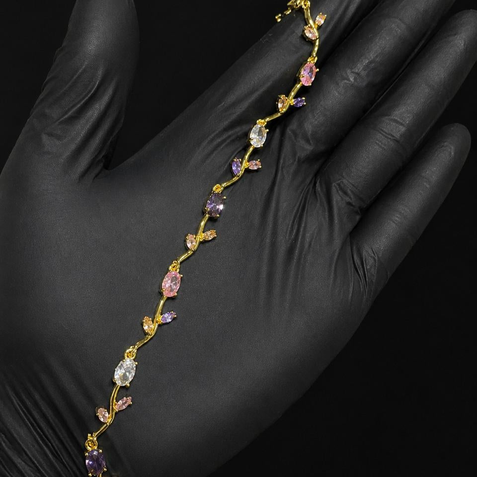 A delicate gold vine-style bracelet with pink, purple, and clear gemstones, displayed on a hand wearing a black glove against a solid black background.
