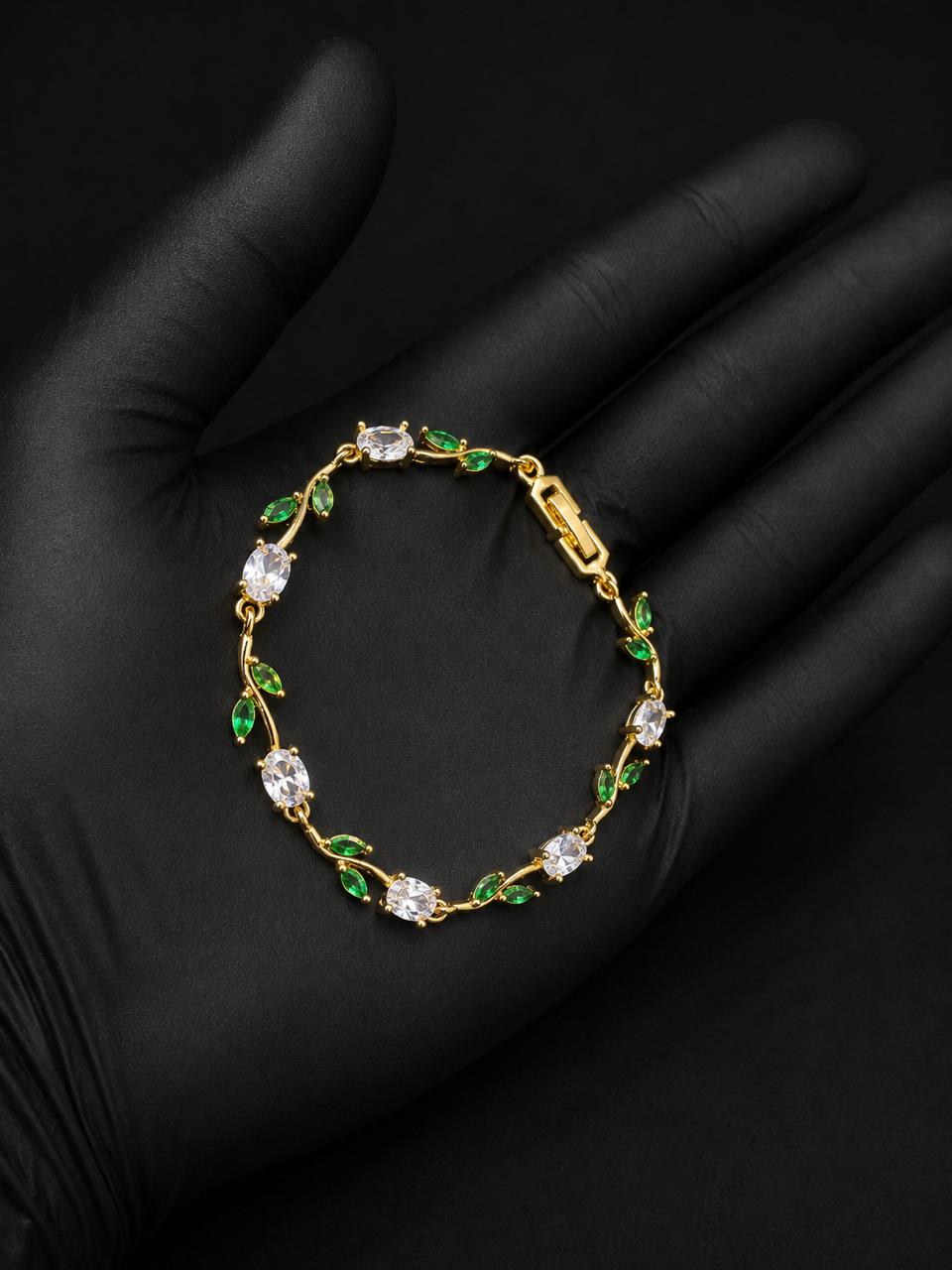A gold bracelet with clear gemstones and green leaf accents, displayed on a black gloved hand.