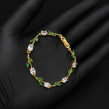 A gold bracelet with clear gemstones and green leaf accents, displayed on a black gloved hand.