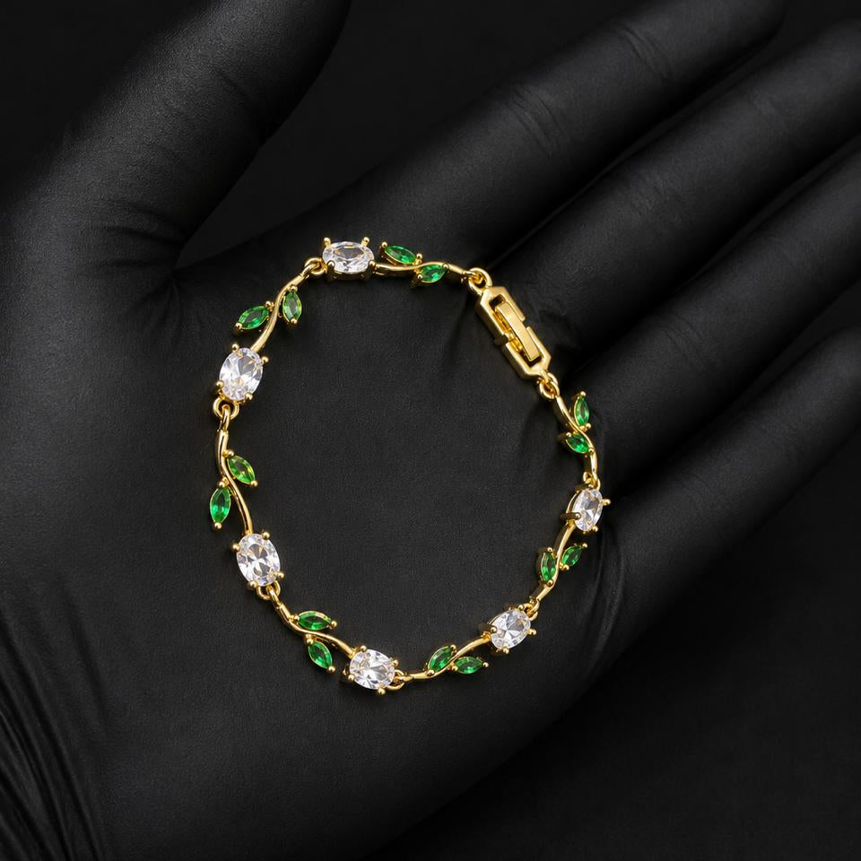 A gold bracelet with clear gemstones and green leaf accents, displayed on a black gloved hand.