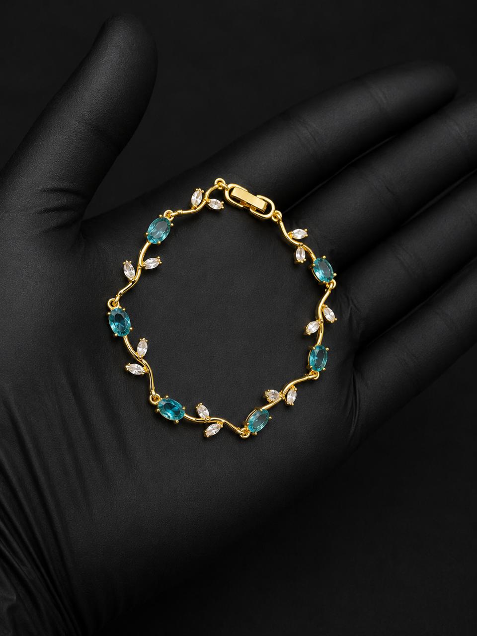 A gold bracelet with marquise-cut blue-green gemstones and small clear stones, displayed on a black gloved hand against a black background.