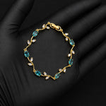 A gold bracelet with marquise-cut blue-green gemstones and small clear stones, displayed on a black gloved hand against a black background.