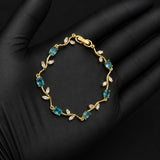 A gold bracelet with marquise-cut blue-green gemstones and small clear stones, displayed on a black gloved hand against a black background.