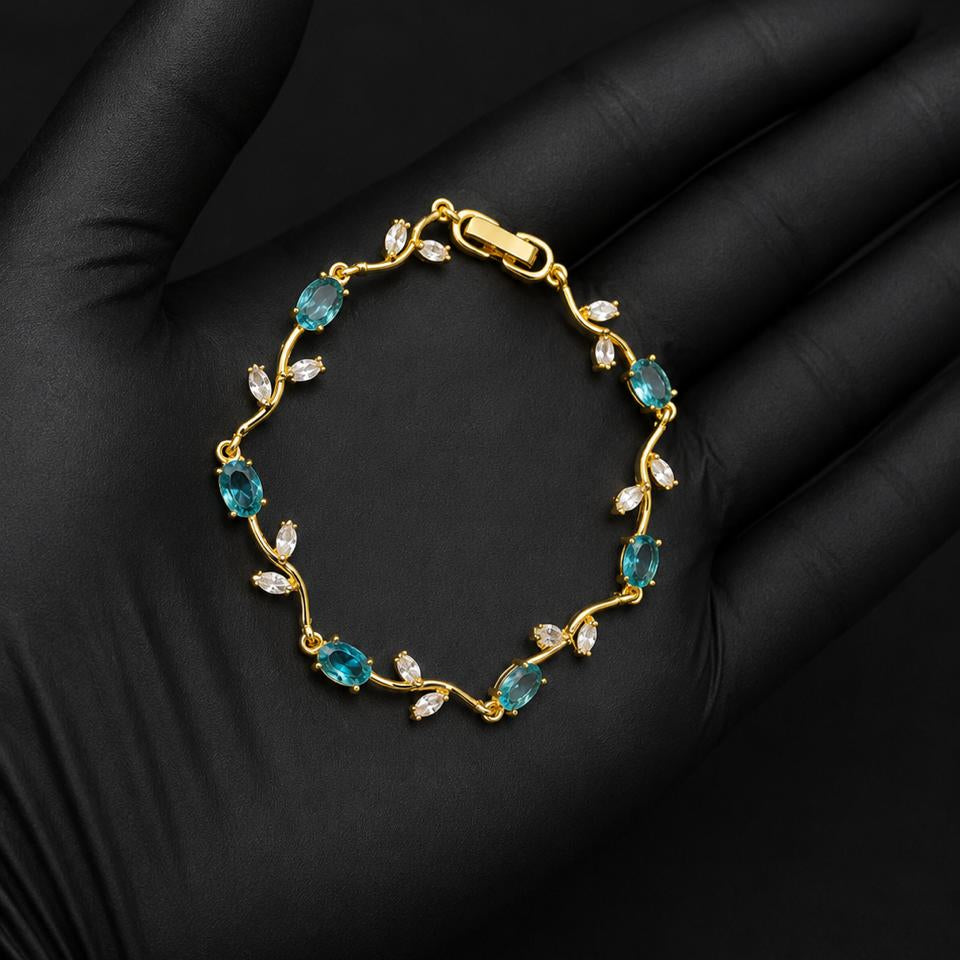 A gold bracelet with marquise-cut blue-green gemstones and small clear stones, displayed on a black gloved hand against a black background.
