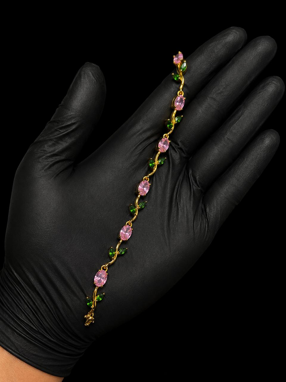 A delicate gold chain bracelet with pink teardrop-shaped crystals and small green leaf accents, held across an open hand wearing a black glove against a solid black background.