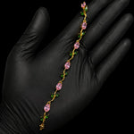A delicate gold chain bracelet with pink teardrop-shaped crystals and small green leaf accents, held across an open hand wearing a black glove against a solid black background.