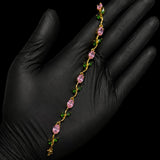 A delicate gold chain bracelet with pink teardrop-shaped crystals and small green leaf accents, held across an open hand wearing a black glove against a solid black background.