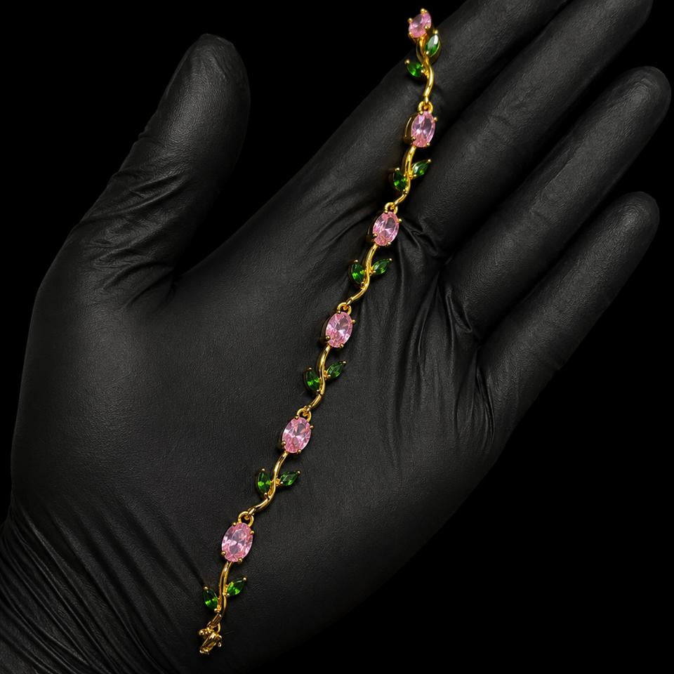 A delicate gold chain bracelet with pink teardrop-shaped crystals and small green leaf accents, held across an open hand wearing a black glove against a solid black background.